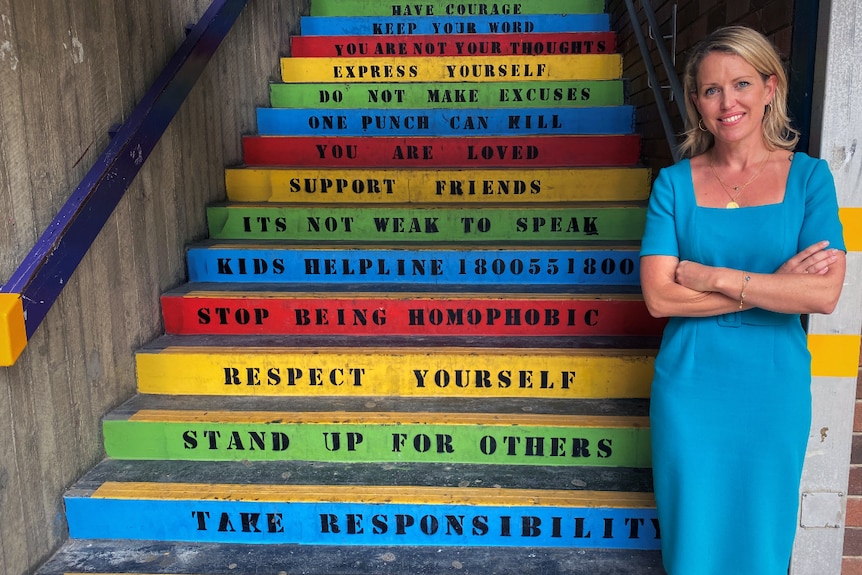 A blonde woman in a blue dress stands next to colourful steps with motivational words painted on them