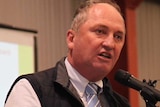 Barnaby Joyce stands on a stage in a recreation centre at a wooden lectern and speaks into a micrphone.