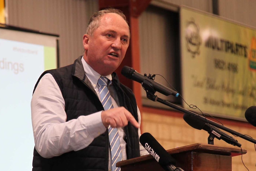 Barnaby Joyce stands on a stage in a recreation centre at a wooden lectern and speaks into a micrphone.