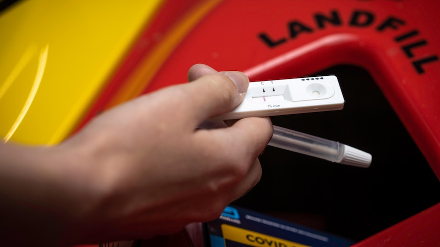 Close up of a hand holding a rapid antigen test above a bin labelled "landfill"