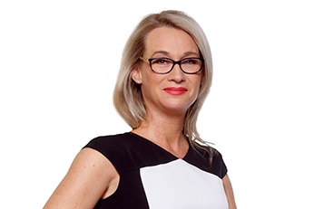 A woman with long blonde hair and black rimmed glasses wears a black and white dress in front of a white backdrop