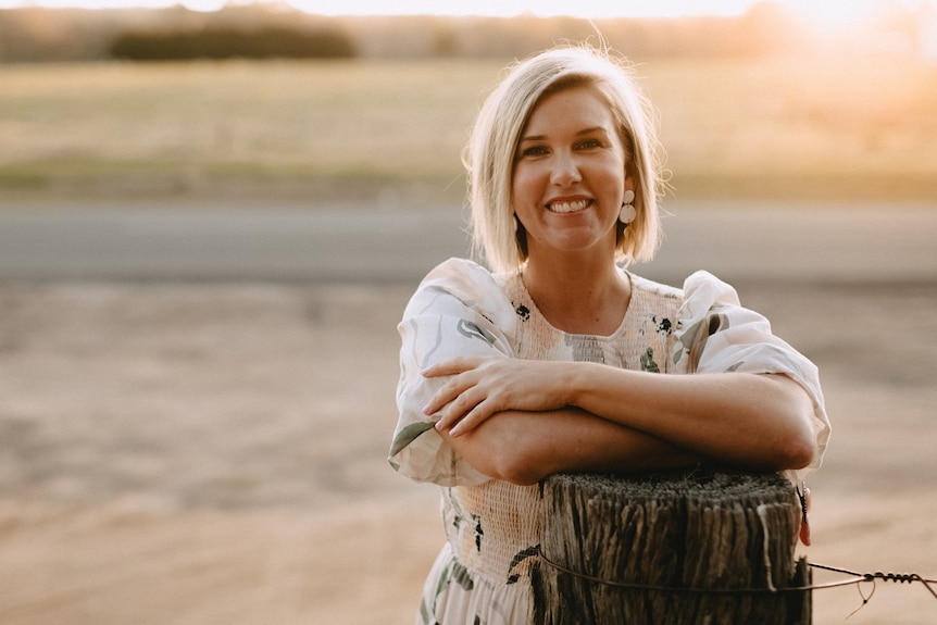 A smiling woman in a dress stands at sunset leans on a fence by a country road.