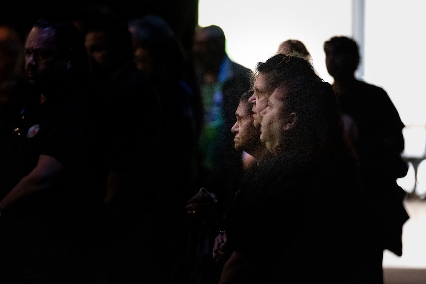A woman walking a dark room in front of a crowd at a funeral service 