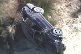 An aerial shot of a dark blue car on its side on a sandy road.