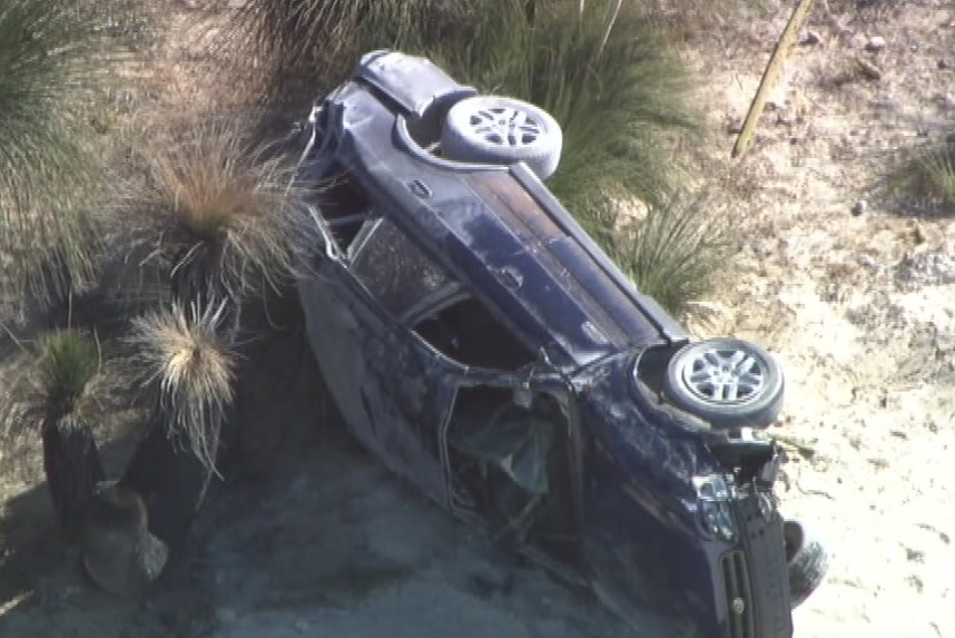 An aerial shot of a dark blue car on its side on a sandy road.