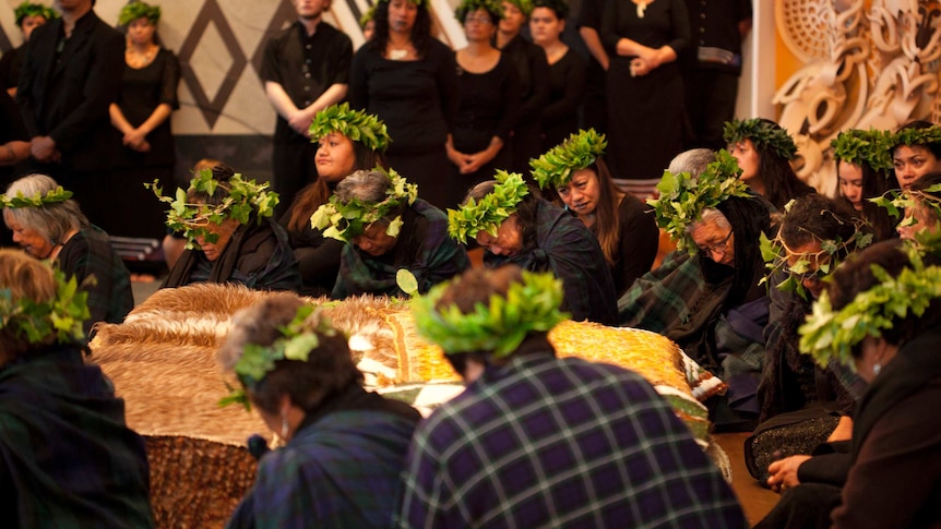 A group of people wearing black and leafy head-wreaths bow in respect around a case containing the remains of an ancestor.