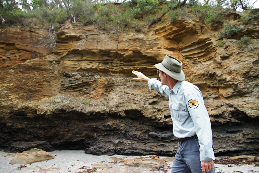 A man in a park ranger's uniform points at a rock formation on a beach.