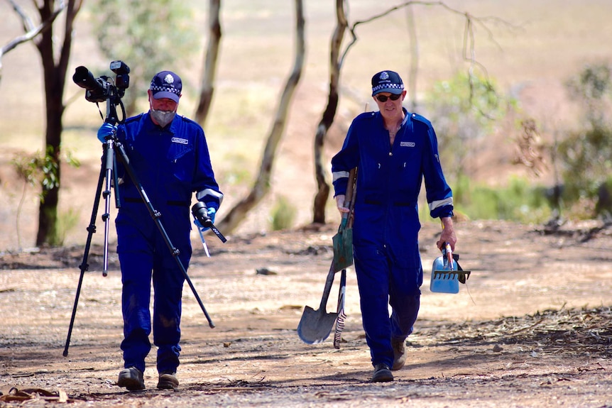 Two forensic police officers carry equipment including a camera, shovel and rake through sparse bushland.