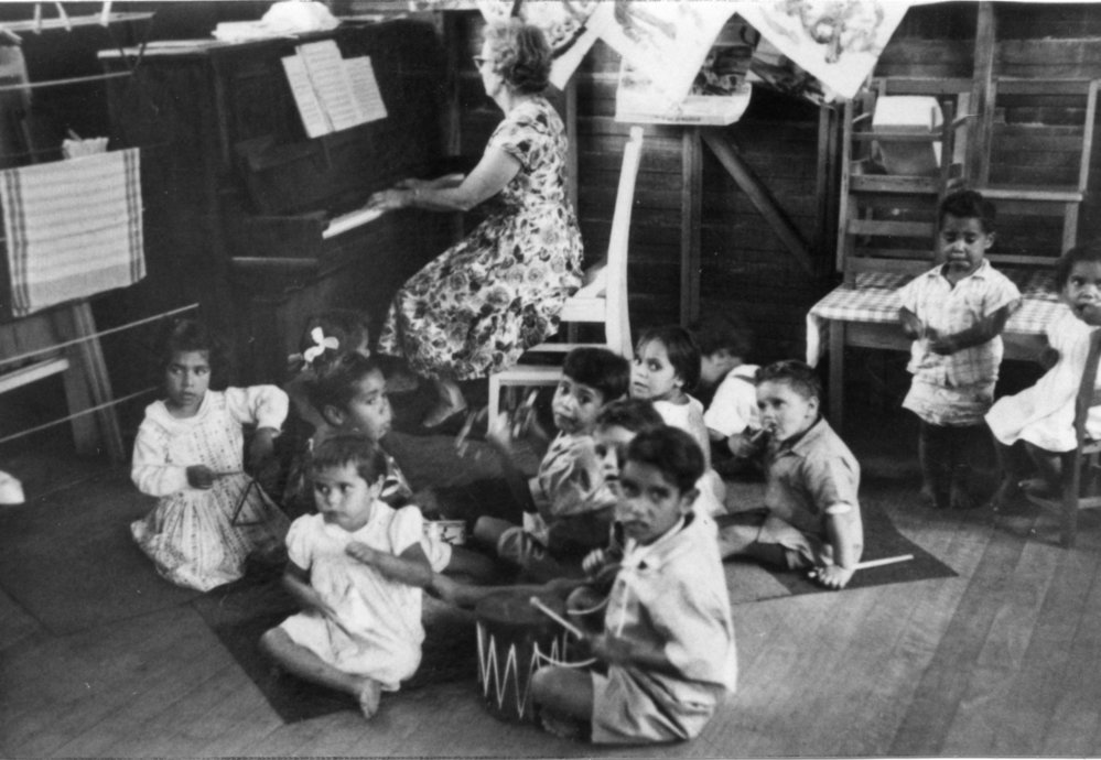 A black and white image of a woman at the piano with 11 Aboriginal children sitting on the floor looking towards the camera