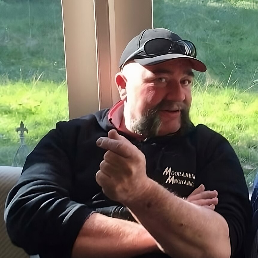 A man with black hat and sunglasses and handlebar mustache holds fingers towards camera, in front of a window.