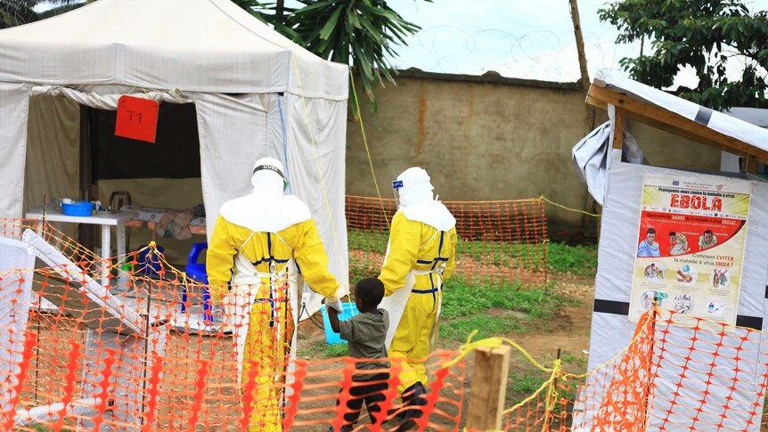 Two health workers in a hazmat suit walk with a small child toward a white medical tent.