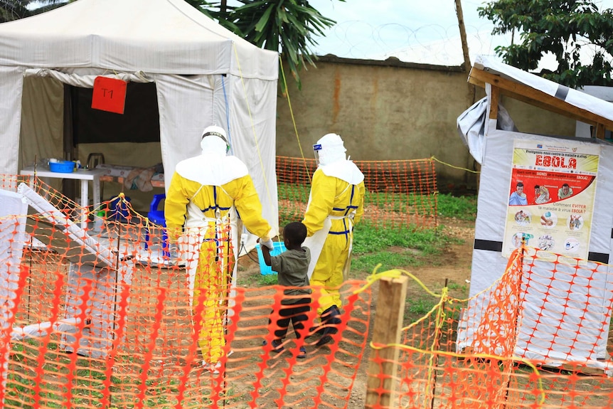 Two health workers in a hazmat suit walk with a small child toward a white medical tent.