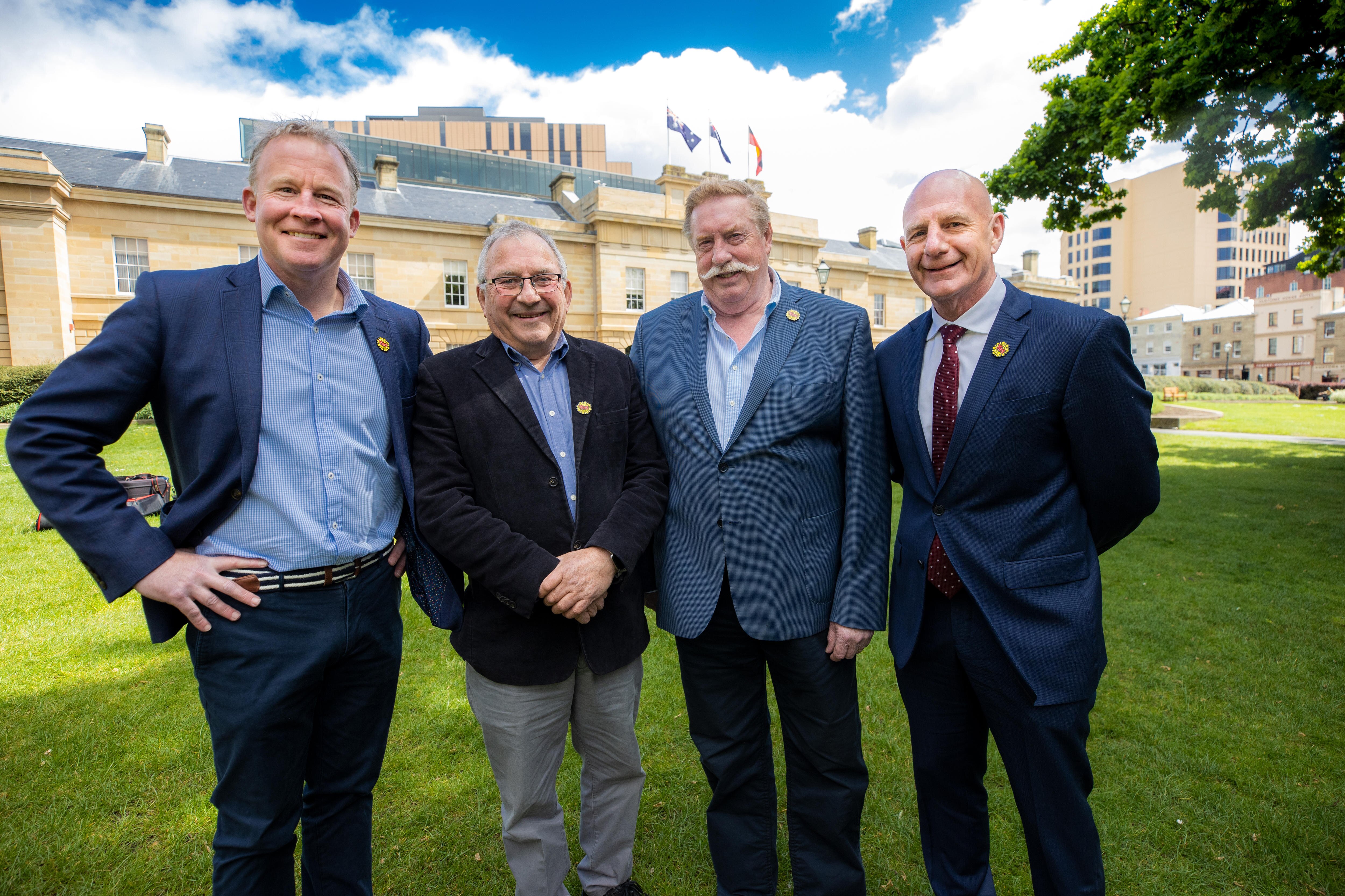 Four men in suits stand in front of a colonial building and smile