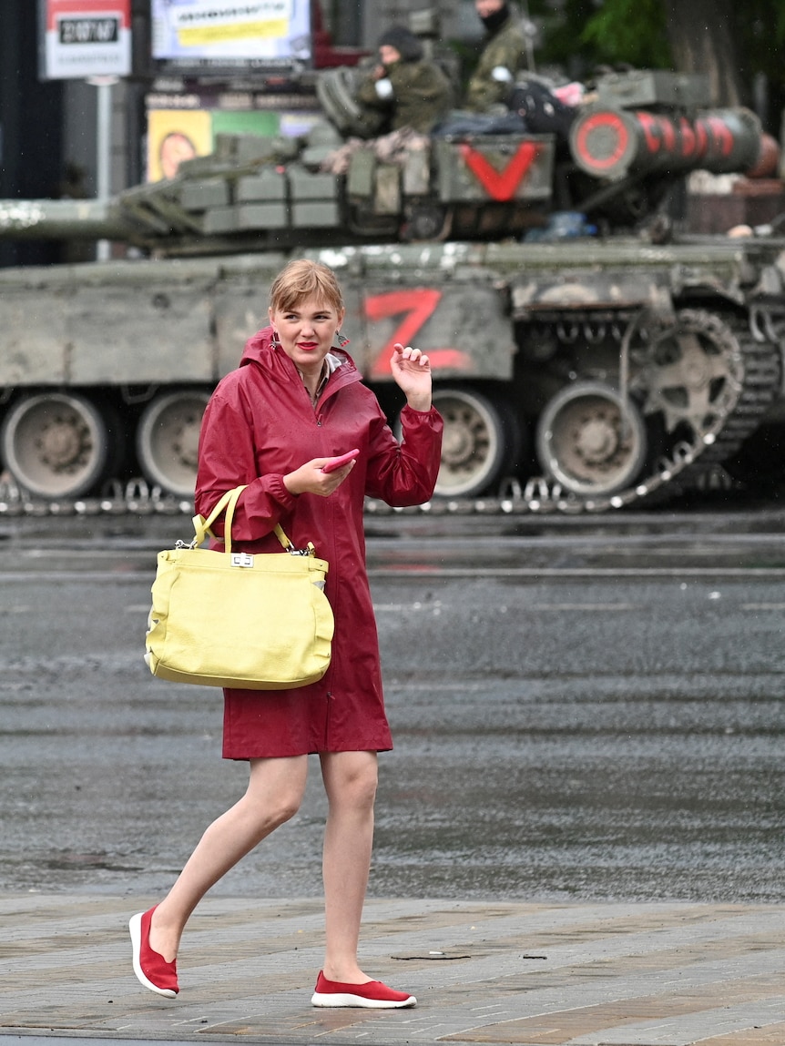 A woman wearing a red coast and holding a yellow bag walks in front of an army tank.