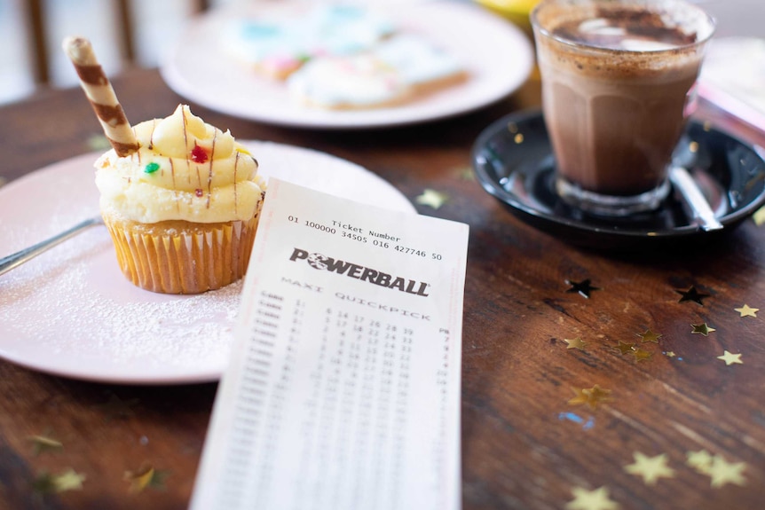 A Powerball lottery ticket resting on a table with a decorated patty cake and a decadent looking coffee.