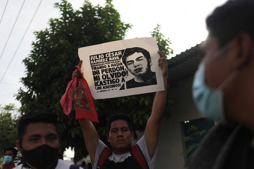 A man holds a poster with a person's face on it and writing in Spanish above his head with both hands