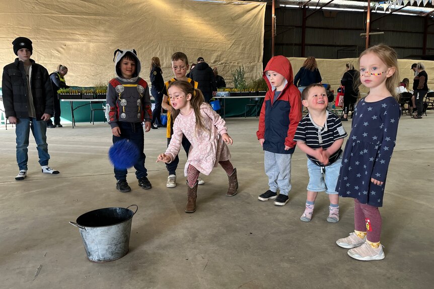 Young kids in a line with a girl in a pink dress throws a blue ball into a metal bucket.
