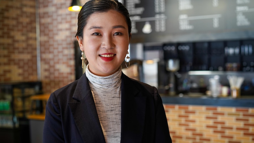 A woman wearing a white turtle neck and black blazer stands smiling in a restaurant.