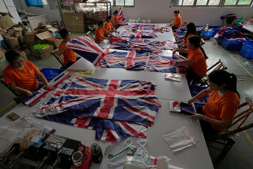 Los trabajadores con camisetas naranjas están sentados en una mesa grande mientras doblan y empaquetan banderas británicas.