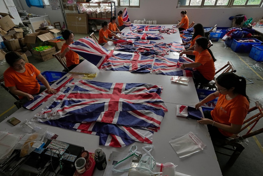 Рабочие в оранжевых рубашках сидят за большим столом и складывают британские флаги.