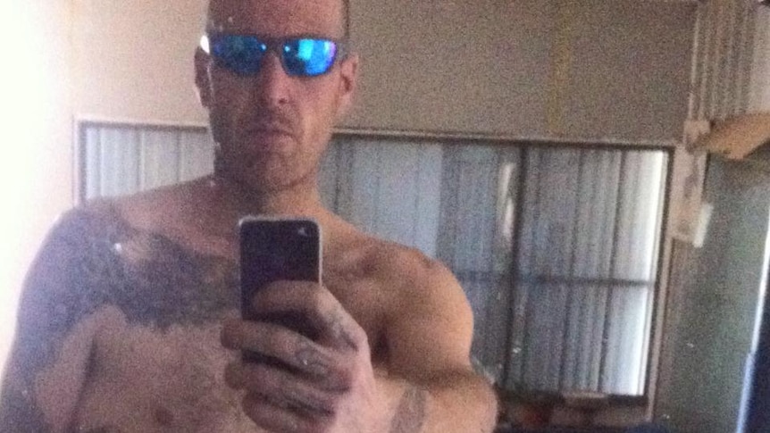 A man in glasses with his shirt off uses a mirror to take a selfie with his phone.