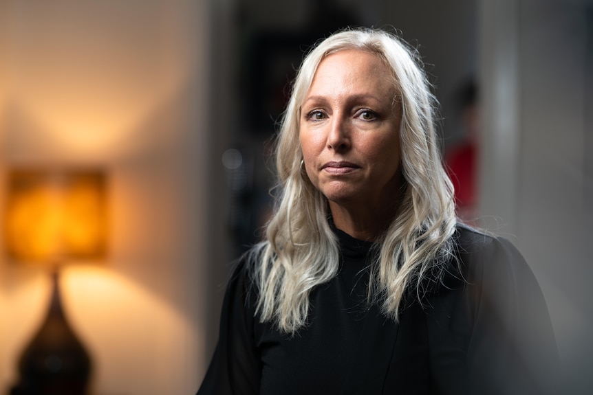 A woman in a black jumper with long white blonde hair looks seriously at the camera. An orange lamp is nearby.