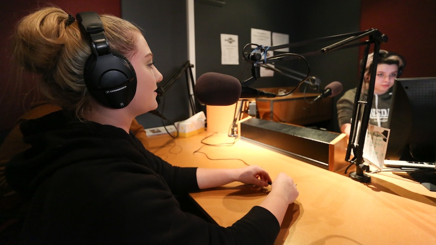 A close up of Jesse behind the microphone wearing headphones with Tiffany in the background looking at the computer screen.