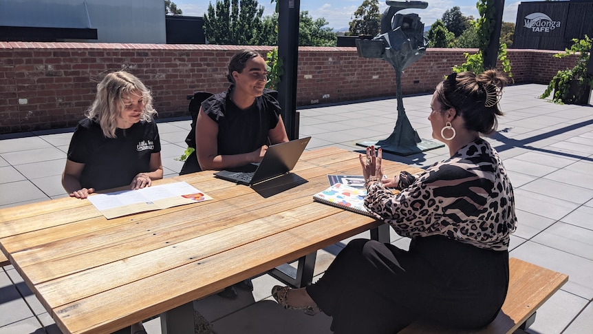 Three women sit talking at a table with a computer and papers.