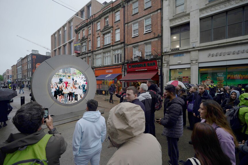 Image of a large grey round portal with a screen in the centre on which people are pictured. Around it are spectators.