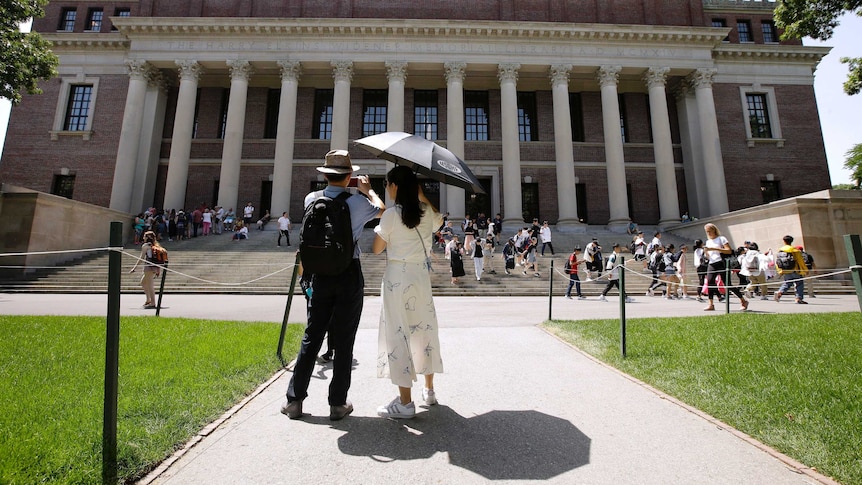 A woman with an umbrella stands next to a man with a camera phone in front of the Harvard library.