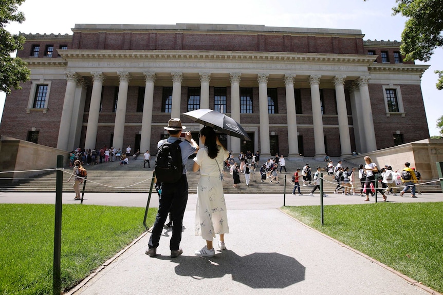 A woman with an umbrella stands next to a man with a camera phone in front of the Harvard library.