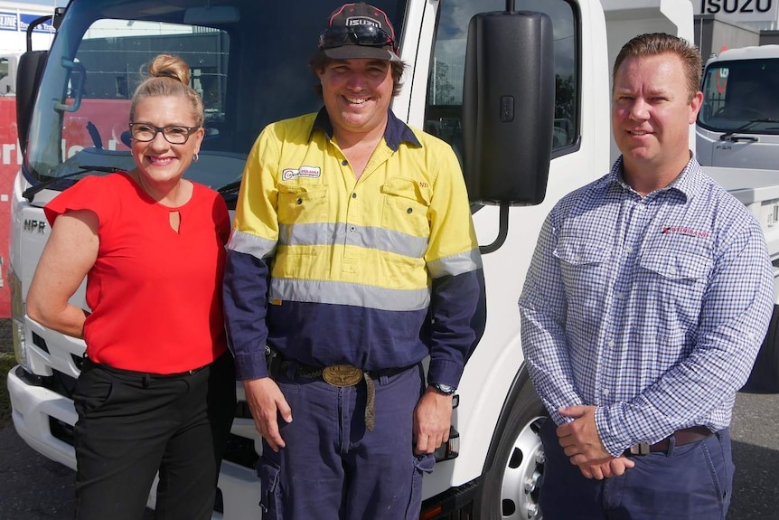 A woman in a red top stands next to a bloke in high-vis and another bloke in a checked shirt in a truck yard