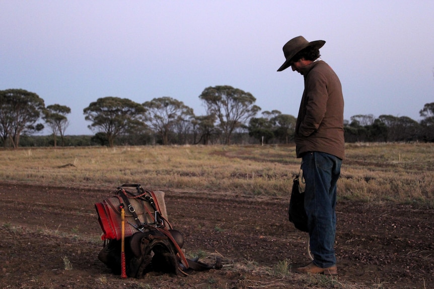 A young man stands in a paddock next to a saddle, looking down at the ground