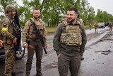 Volodymyr Zelenskyy stands in a road wearing a flak jacket with several other soldiers. 
