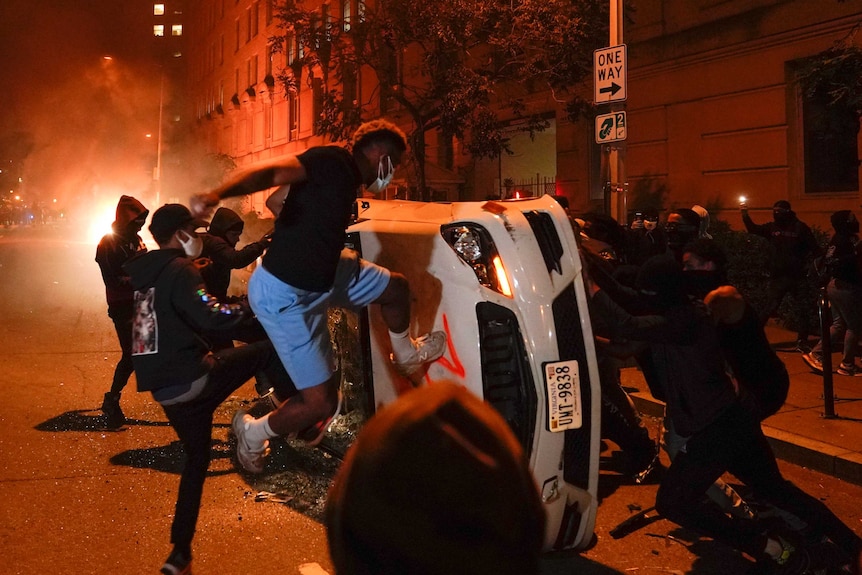 A group of young men wearing face masks kick a car that has been pushed onto its side while fires burn in the background.