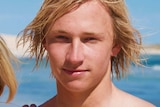 A head shot showing a young man with longer blond hair posing for a photo at the beach.