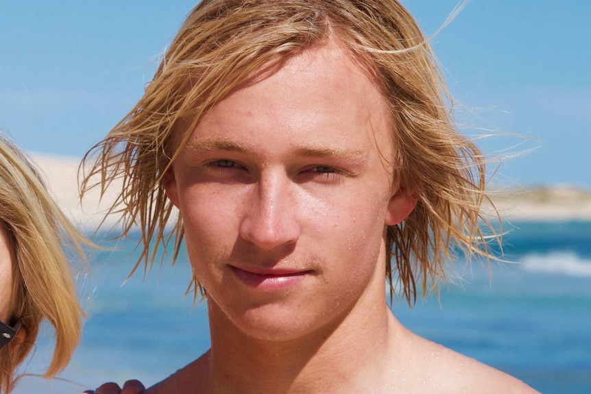 A head shot showing a young man with longer blond hair posing for a photo at the beach.