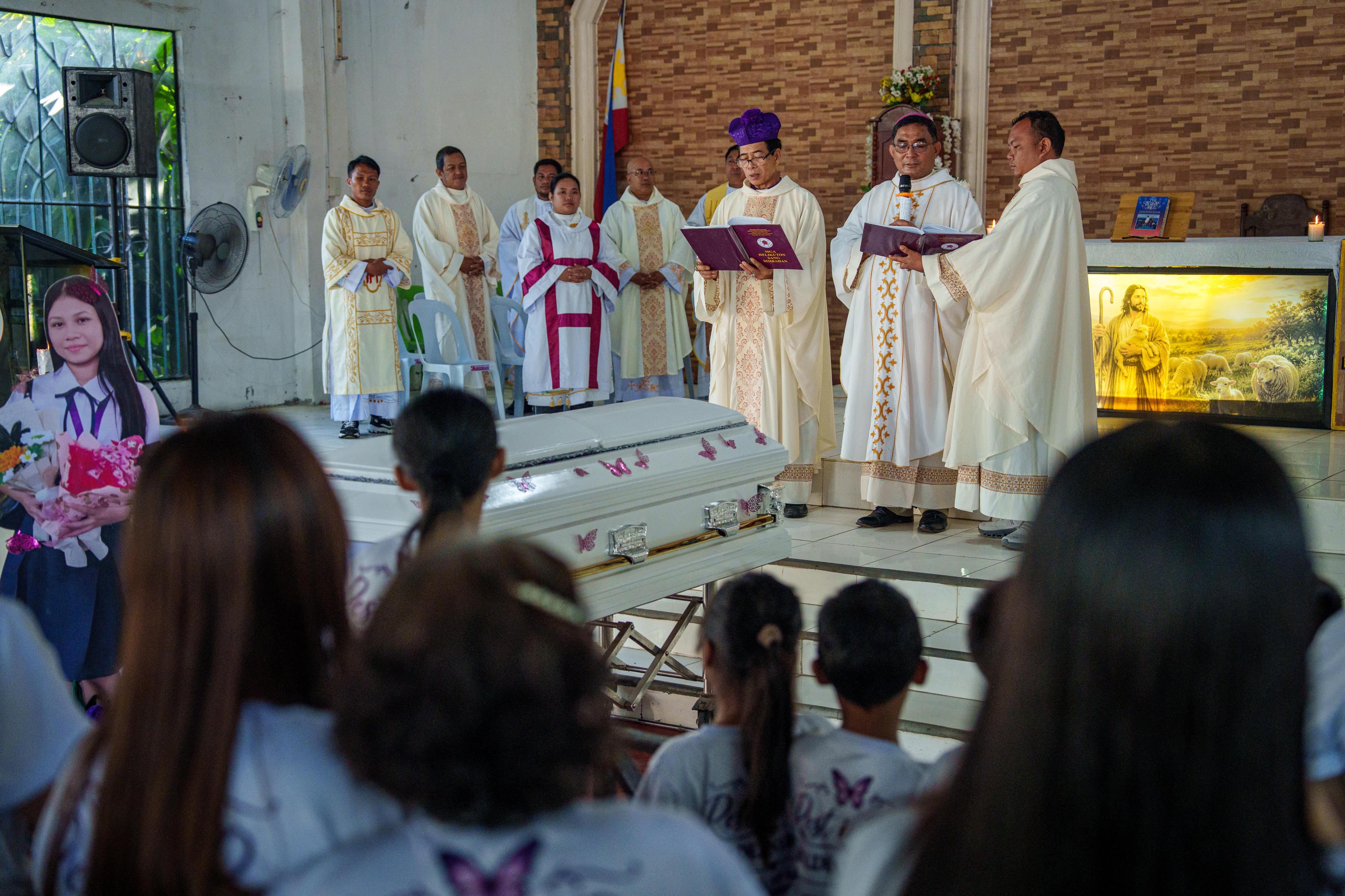 Church ministers conduct a funeral service in a light coloured church with a white coffin.