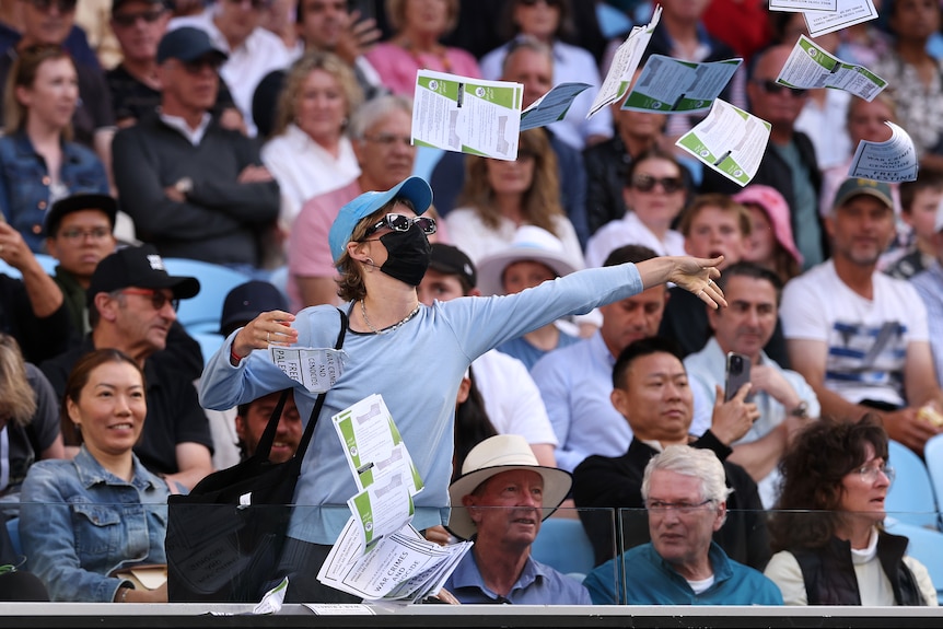 A woman, wearing a hat and face mask, throws paper from a grandstand onto a tennis court
