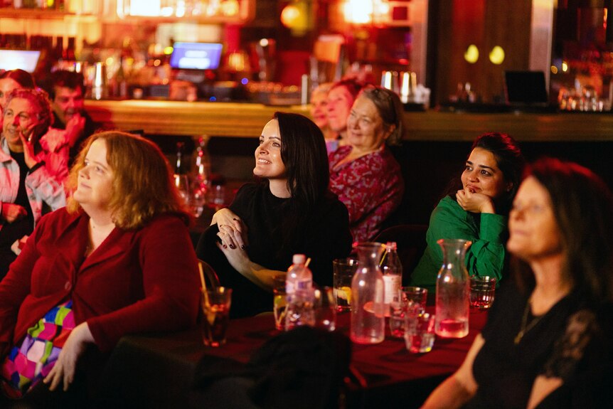 An audience of people sitting in a pub looking towards a stage, looking amused and happy, drinks on table