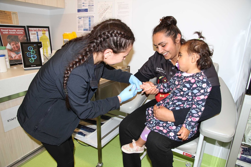 A nurse wears blue gloves as she pricks a young girl's finger with a small needle while the girl sits on her smiling mum's lap