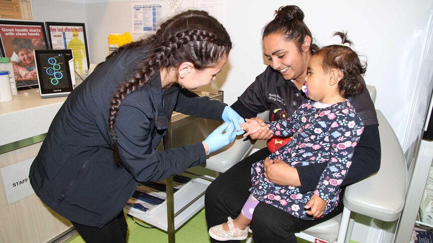 A nurse wears blue gloves as she pricks a young girl's finger with a small needle while the girl sits on her smiling mum's lap