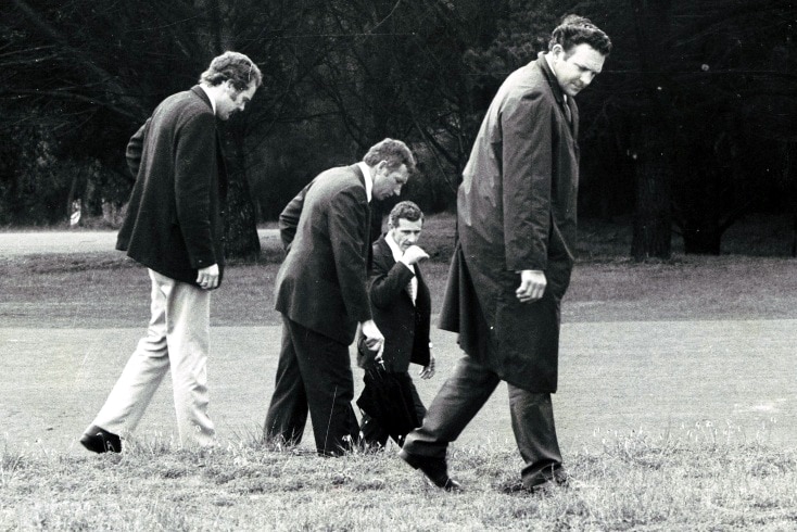 A black and white photo of four men with their heads down looking at the grass