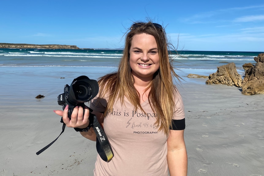 Woman standing with camera in hand smiling at camera at beach, bluff and ocean in the background.