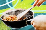 A person holds a steaming frypan, stirring it with a wooden spoon, making a light stir fry for dinner.