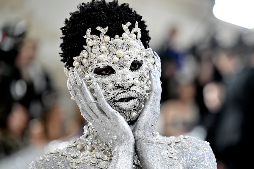 Close up of Lil Nas X whose face is covered in silver jewels and pearls.