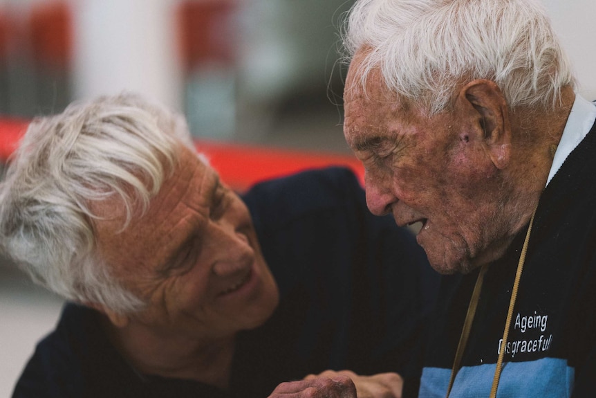 A man with grey hair and a black T-shirt kneels down and talks to an elderly man in a wheelchair.