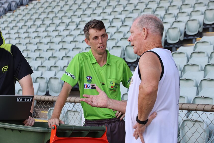 Umpire Sam Cunningham wearing his grey and green NTFL umpiring uniform talks to a man in the grandstand.