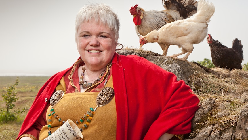 Icelandic priestess dressed in traditional clothing, holding a bull horn, with chickens behind her.