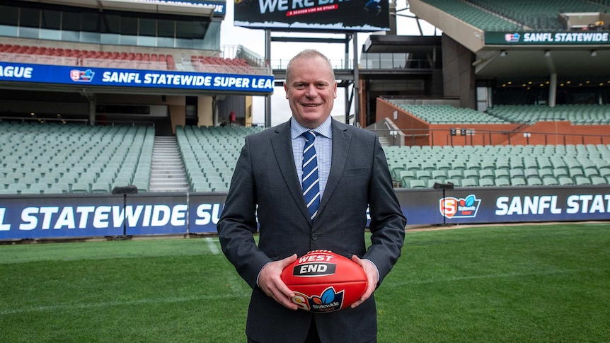 A man in a suit stands before an empty oval grandstand with the words We're Back on display behind him.